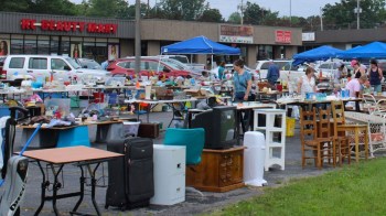 A bustling outdoor flea market with various items for sale and people browsing tables under blue tents.