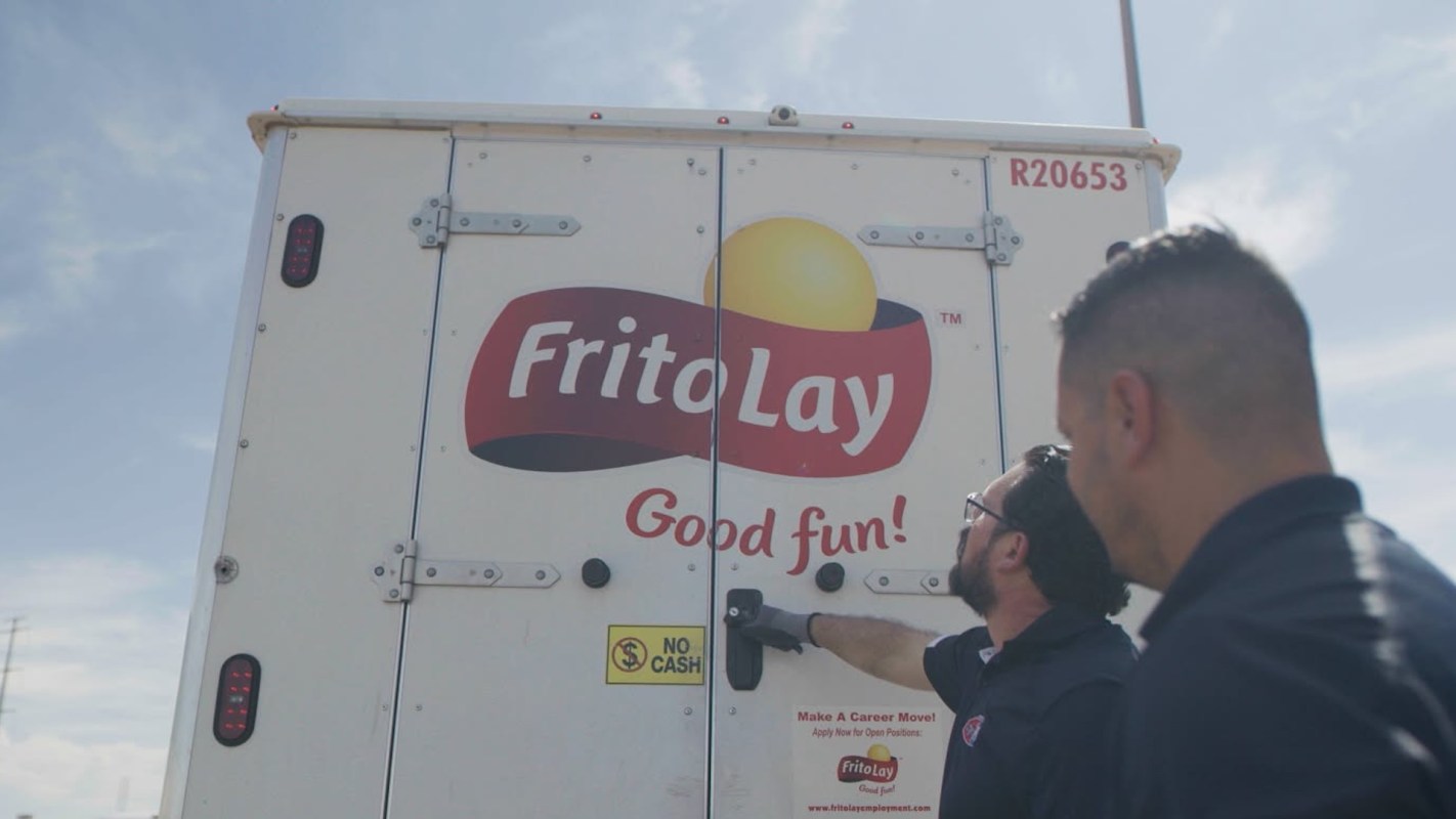 Two men are examining the back of a Frito-Lay delivery truck under a clear blue sky.