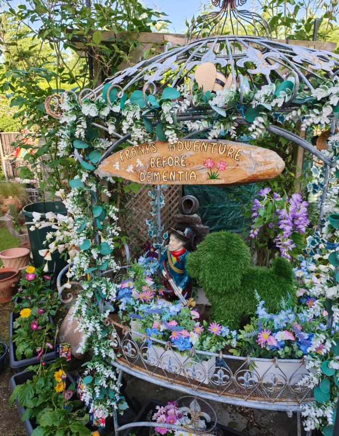 A whimsical garden display featuring a statue, a faux dog, and flowers beneath a sign reading "Frank's Adventure Before Dementia."