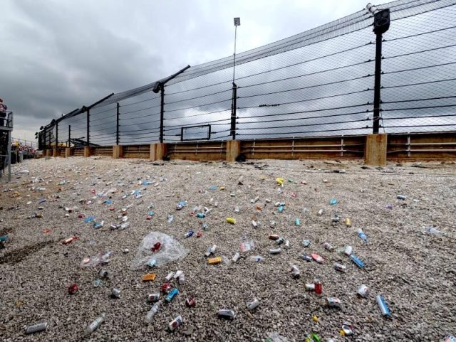 A raceway fence overlooking a litter-strewn area under a cloudy sky, with spectators visible in the background.