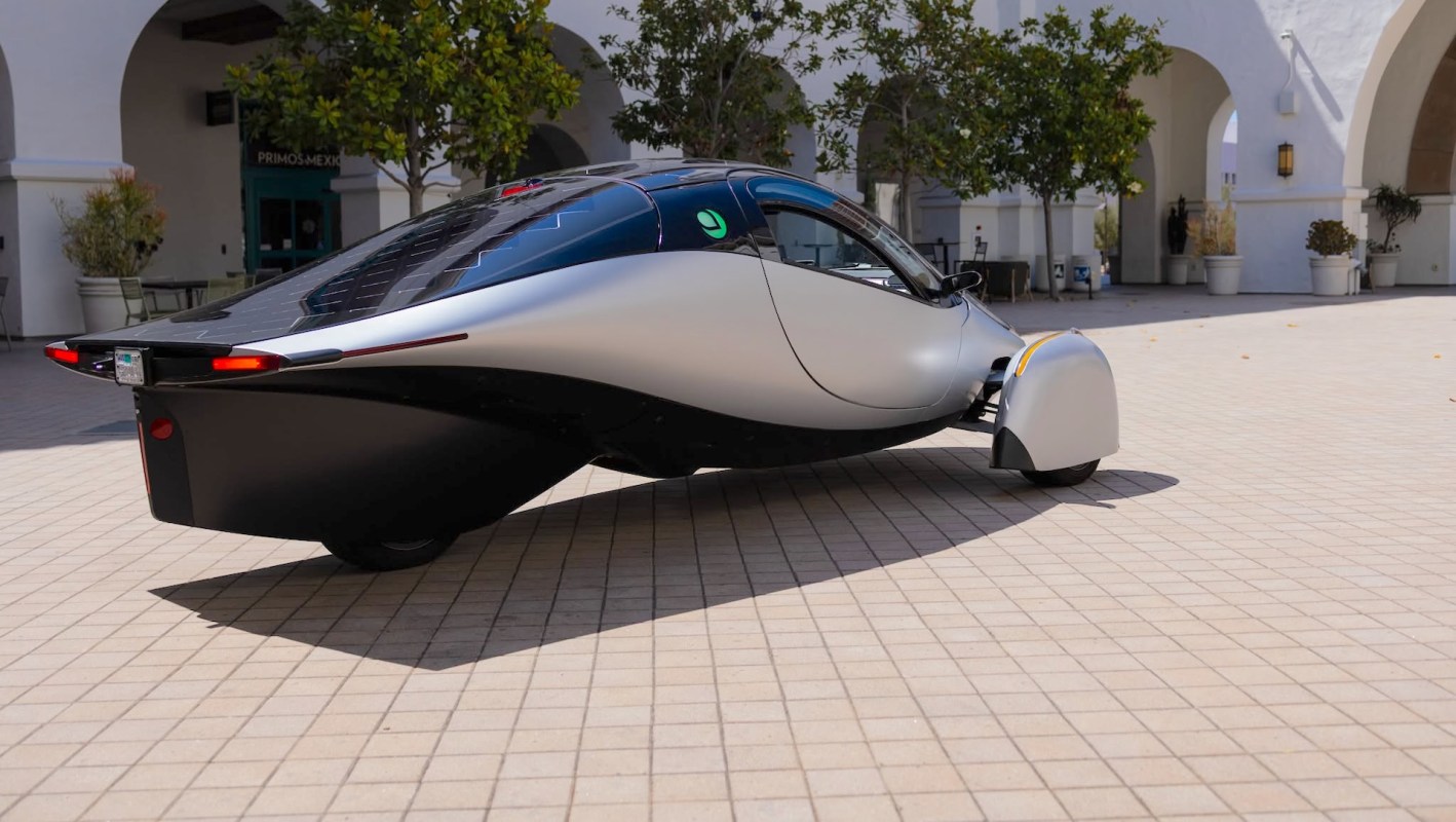 A three-wheeled electric vehicle with solar panels on top parked on a tiled surface with green trees in the background.