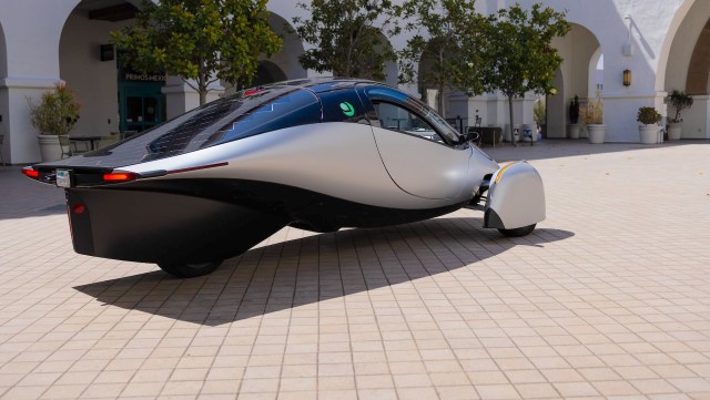 A three-wheeled electric vehicle with solar panels on top parked on a tiled surface with green trees in the background.