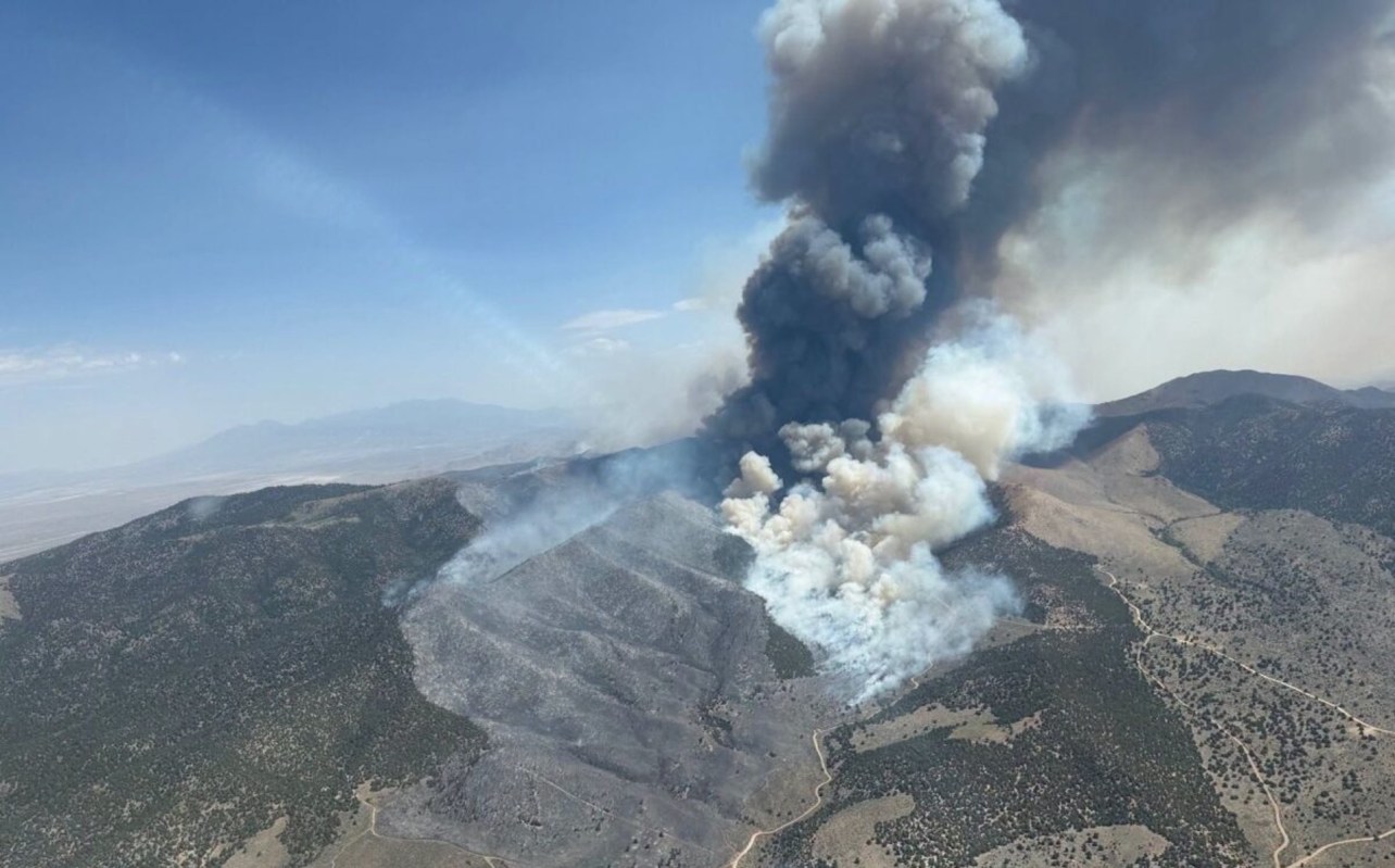 A large plume of dark smoke rises from a wildfire in mountainous terrain.