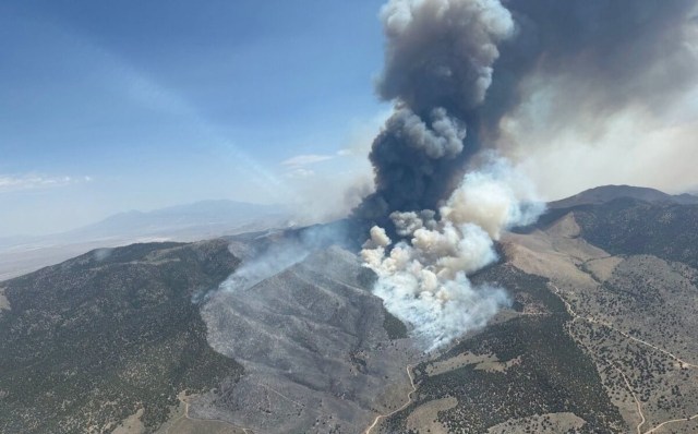 A large plume of dark smoke rises from a wildfire in mountainous terrain.