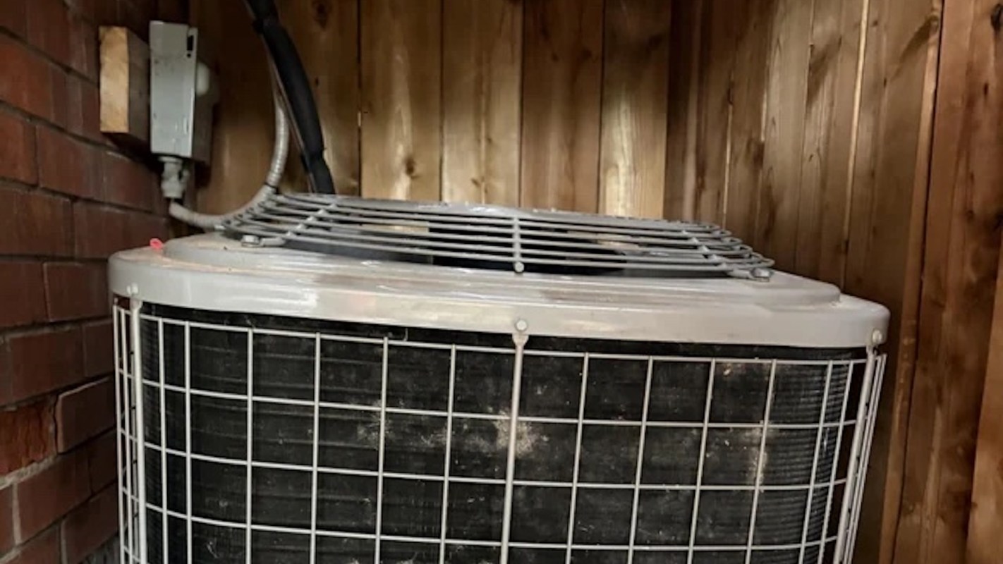 An outdoor air conditioning unit placed against a wooden wall and adjacent to a brick structure.