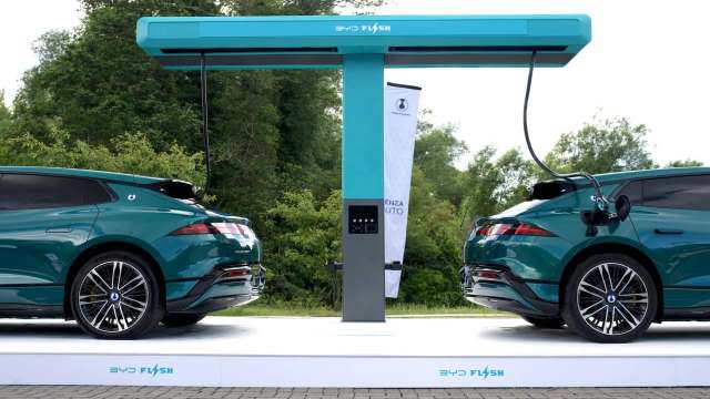 Two teal electric cars parked at a charging station with the words "BYD Flash" on it, surrounded by greenery.