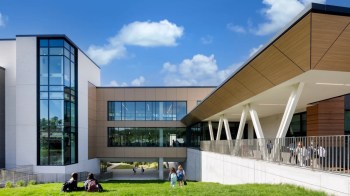 A modern educational building surrounded by green grass and clear blue skies, with students walking and sitting nearby.