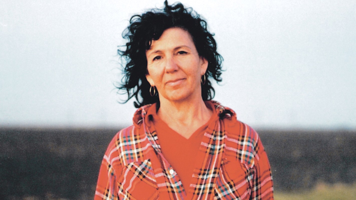 A woman with curly hair wearing a red plaid shirt stands against a soft, blurred background.