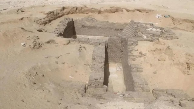Ruins of an ancient structure partially excavated in a sandy desert environment.