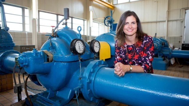 A woman in a floral dress poses with industrial blue pumps and gauges in a bright, spacious facility.