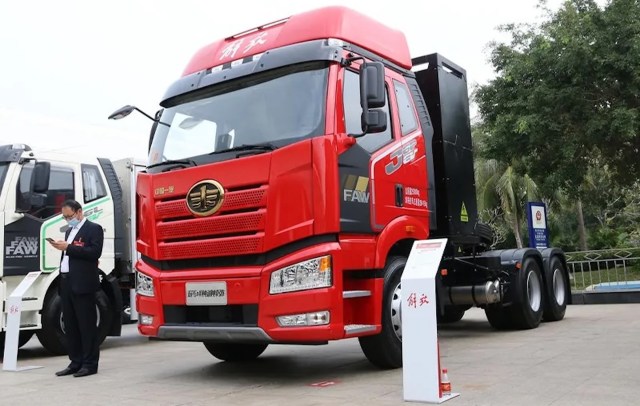 A red FAW truck is displayed at an exhibition with a man in a suit checking his phone nearby.