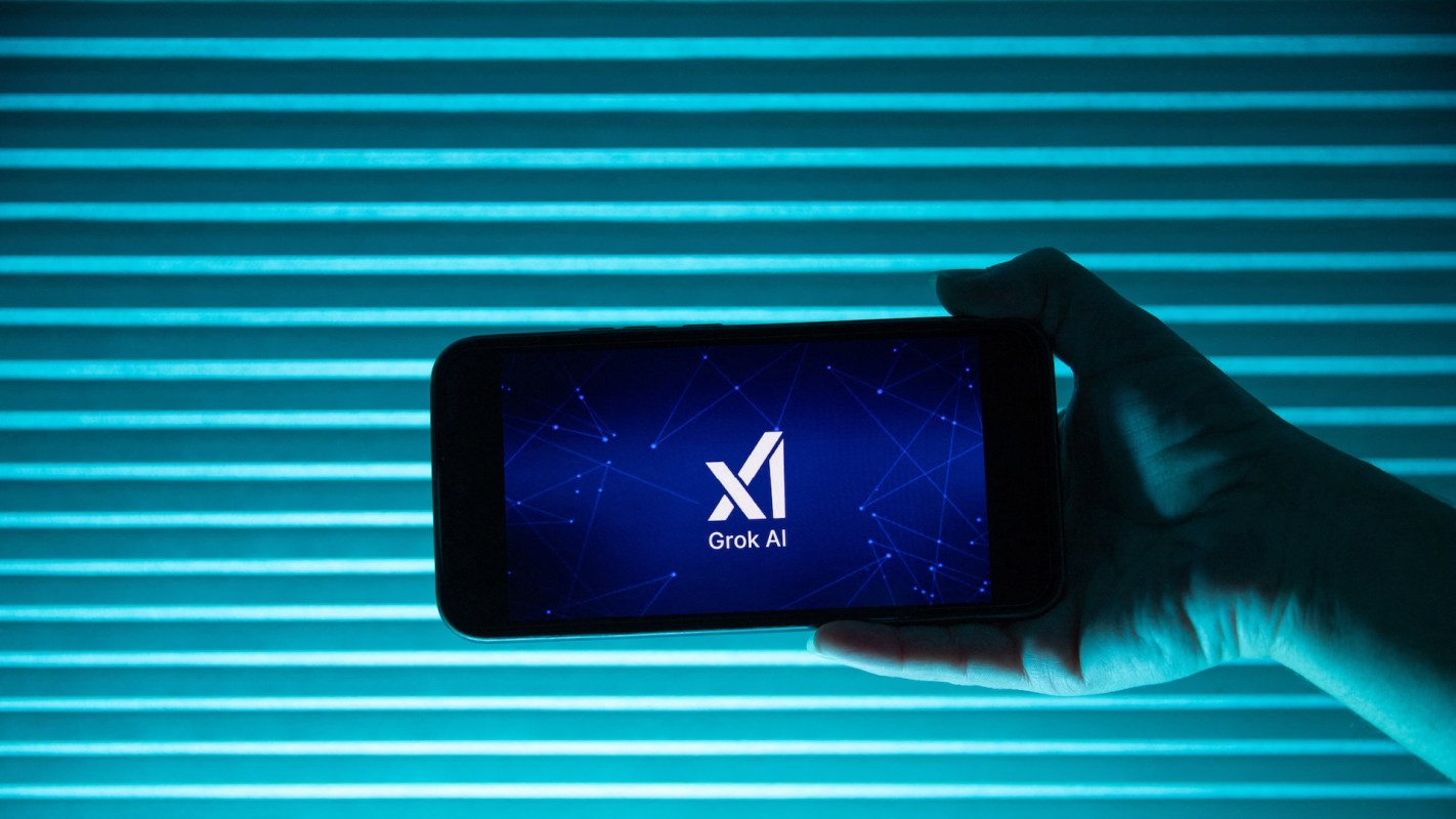 A hand holds a smartphone displaying the "Grok AI" logo against a teal striped background.