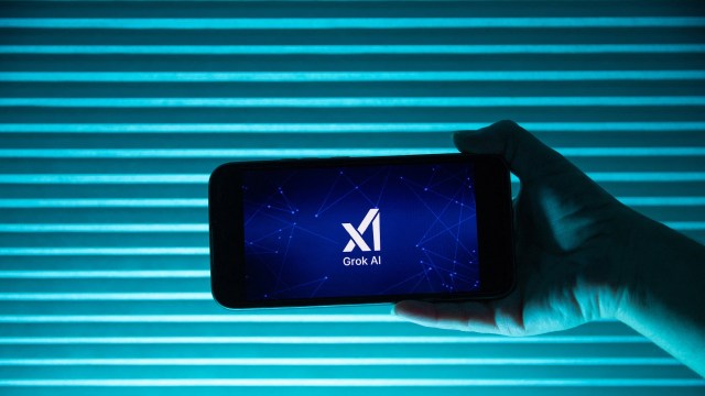 A hand holds a smartphone displaying the "Grok AI" logo against a teal striped background.