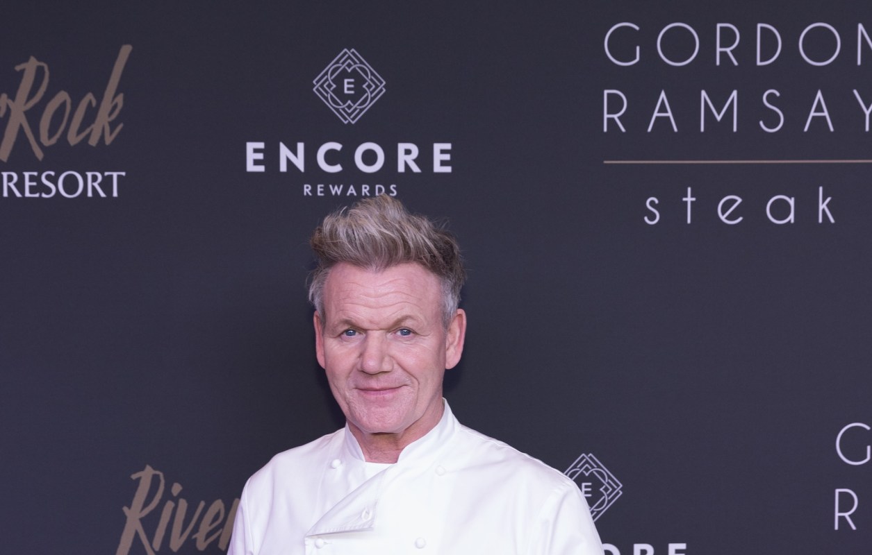 Celebrity chef Gordon Ramsay, in a white coat, stands in front of a sign for Gordon Ramsay Steak.