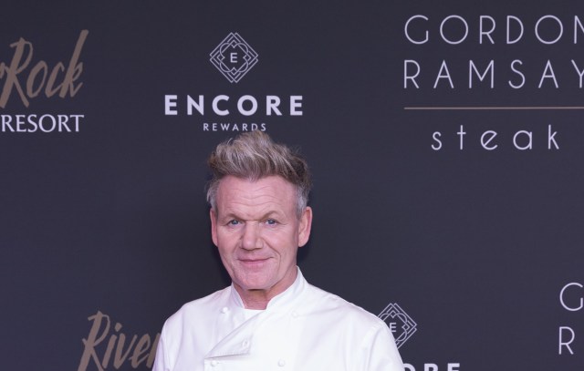Celebrity chef Gordon Ramsay, in a white coat, stands in front of a sign for Gordon Ramsay Steak.
