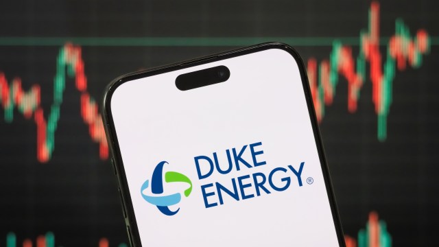 A smartphone displaying the Duke Energy logo in front of a blurred stock market graph.