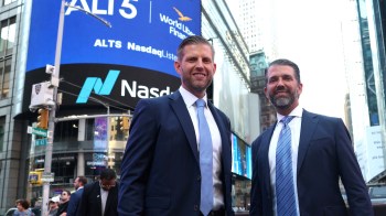 Eric Trump and Donald Trump Jr. in suits pose in Times Square, with a large Nasdaq sign in the background.