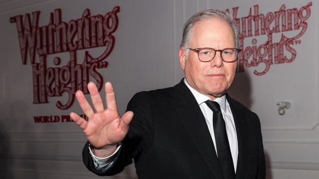 Warner Bros. CEO David Zaslav, in a suit, waves his hand at a Wuthering Heights world premiere event backdrop.