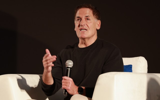 A man in a black shirt, Mark Cuban, speaks into a microphone while gesturing, seated in a white chair.