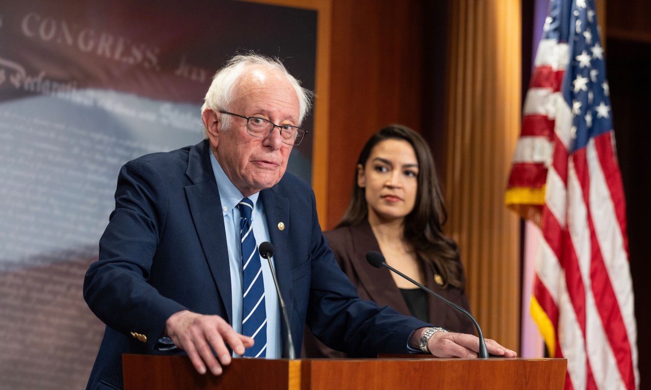 Bernie Sanders speaks at a podium with Alexandria Ocasio-Cortez standing behind him, an American flag in the background.