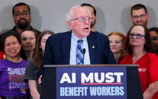 Bernie Sanders, with glasses, speaks at a podium with a sign that reads "AI must benefit workers."