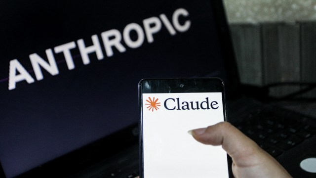 A person holding a smartphone displaying the "Claude" logo, with the word "ANTHROPIC" in the background on a laptop screen.