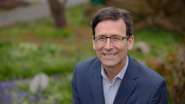 Washington Governor Bob Ferguson, in a suit, poses outdoors with a blurred garden background.