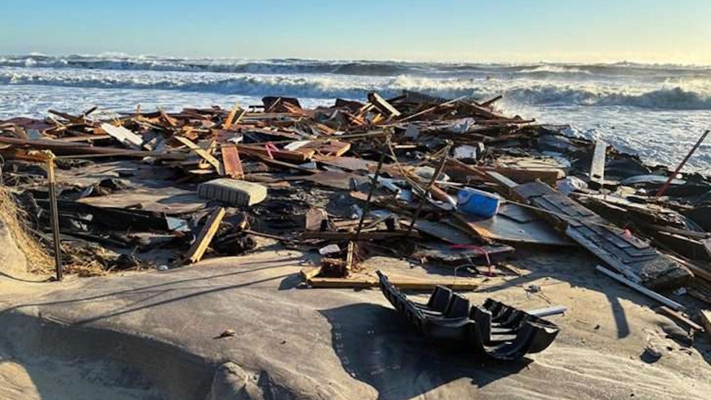 Debris from a storm, including wood and objects, scattered on a beach near crashing waves.