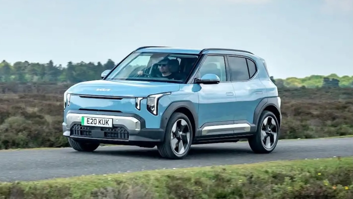 A blue Kia electric SUV drives along a rural road with trees and greenery in the background.