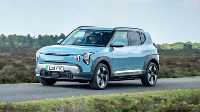 A blue Kia electric SUV drives along a rural road with trees and greenery in the background.