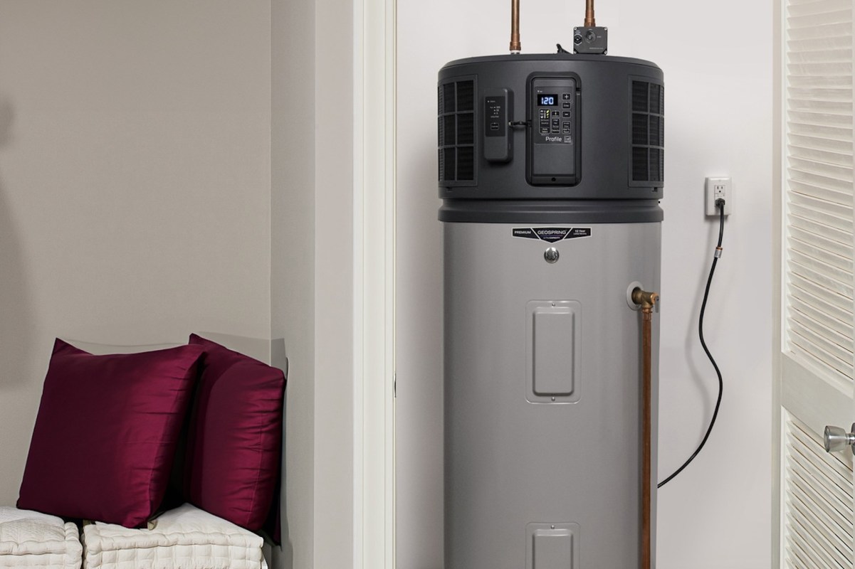 A gray heat pump water heater stands beside burgundy cushions in a modern room with light-colored walls.