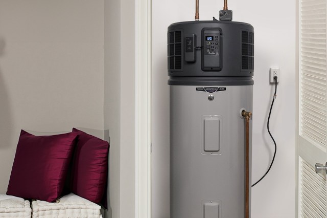 A gray heat pump water heater stands beside burgundy cushions in a modern room with light-colored walls.