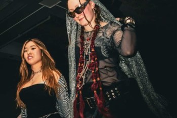 Two young women walk together, one wearing sunglasses and layered jewelry, while the other sports a black dress.