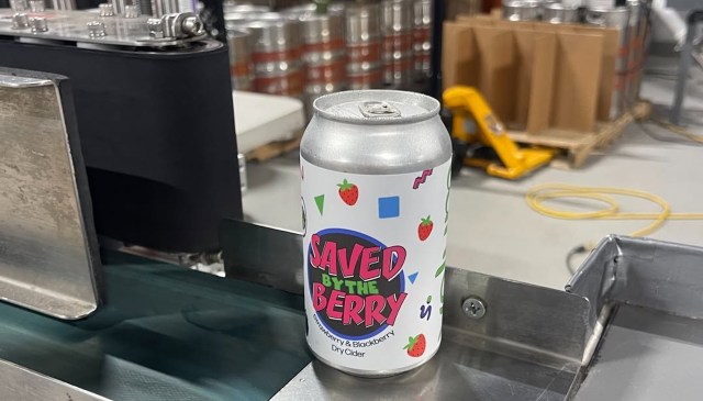 A can of "Saved By The Berry" cider with a colorful design sits on a conveyor in a brewery.