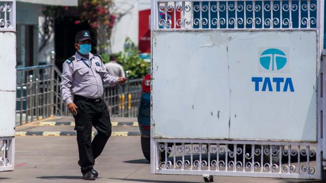 A security guard in uniform and mask walks past a gated entrance with the TATA logo.