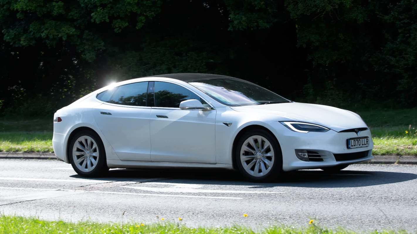 A white Tesla Model S is driving along a road lined by grass.