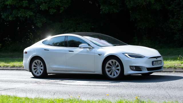 A white Tesla Model S is driving along a road lined by grass.