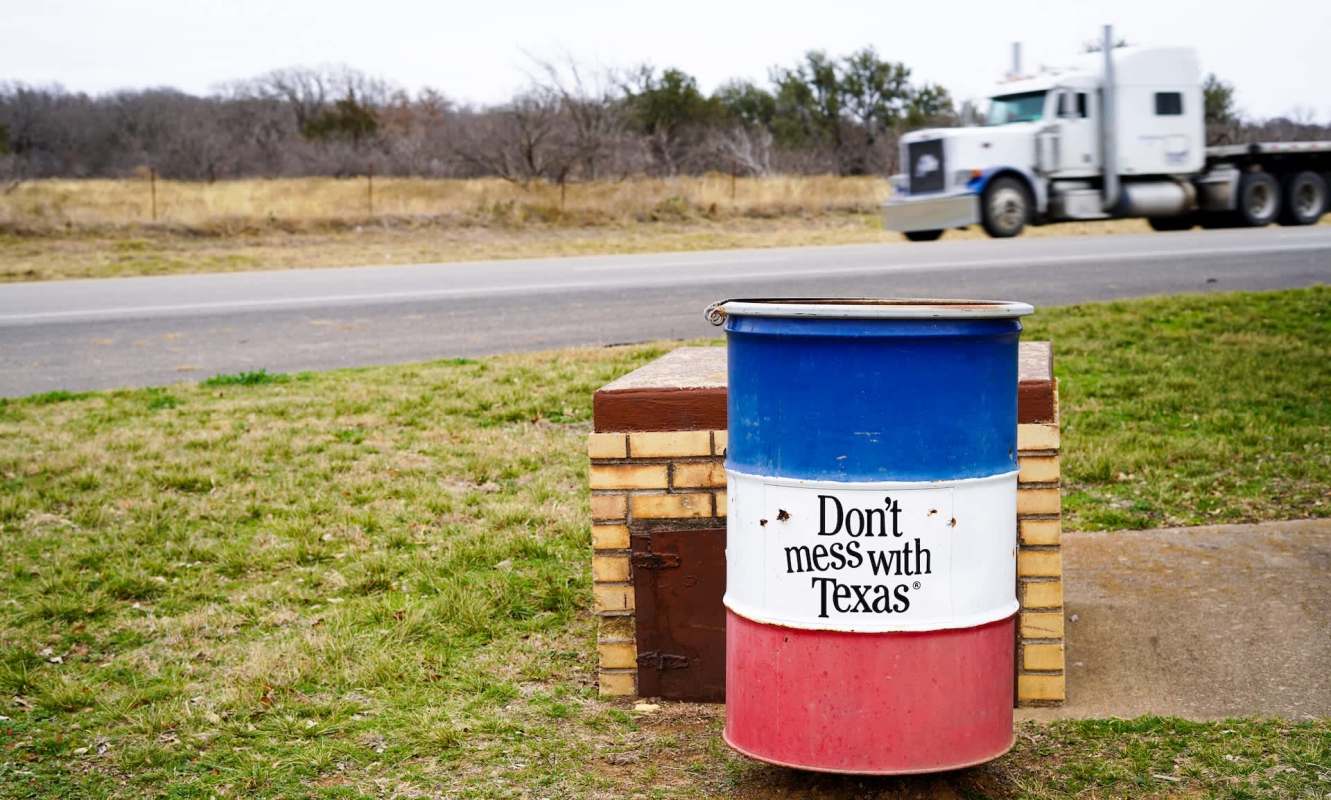 A colorful trash barrel with a "Don't mess with Texas" sign beside a roadside, with a truck passing in the background.