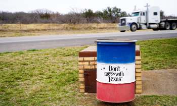 A colorful trash barrel with a "Don't mess with Texas" sign beside a roadside, with a truck passing in the background.