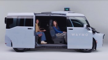 Two women smile while sitting inside a blue Waymo autonomous vehicle with an open sliding door.