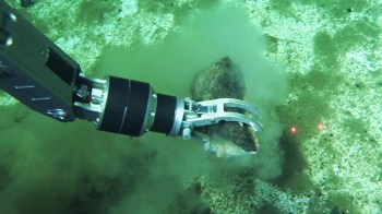 A robotic arm digs into the seabed, stirring up sediment while grasping a rock.
