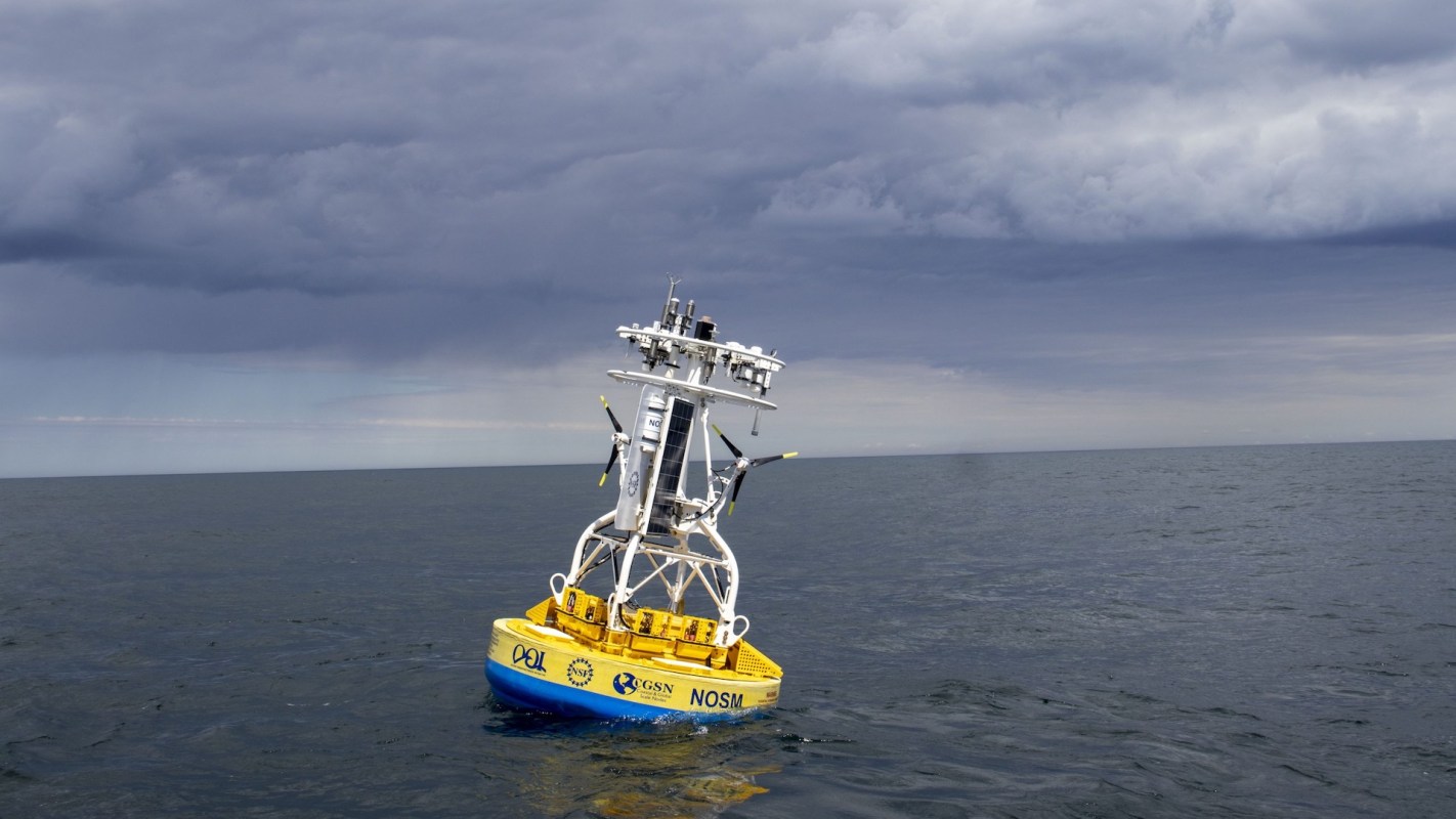 A yellow ocean buoy equipped with sensors in calm waters under a cloudy sky.