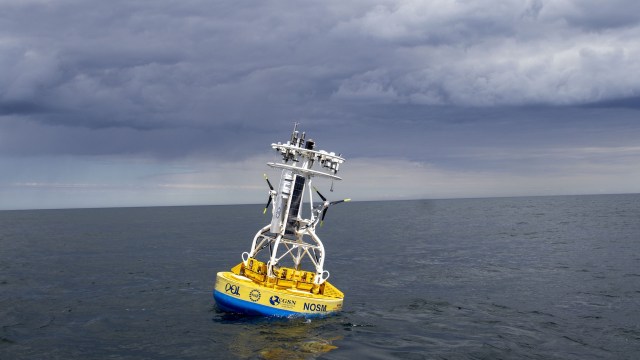 A yellow ocean buoy equipped with sensors in calm waters under a cloudy sky.