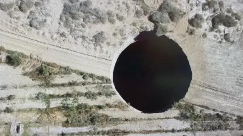 A top-down look at a large hole in that covers the width of a dirt track and surrounding grassland.