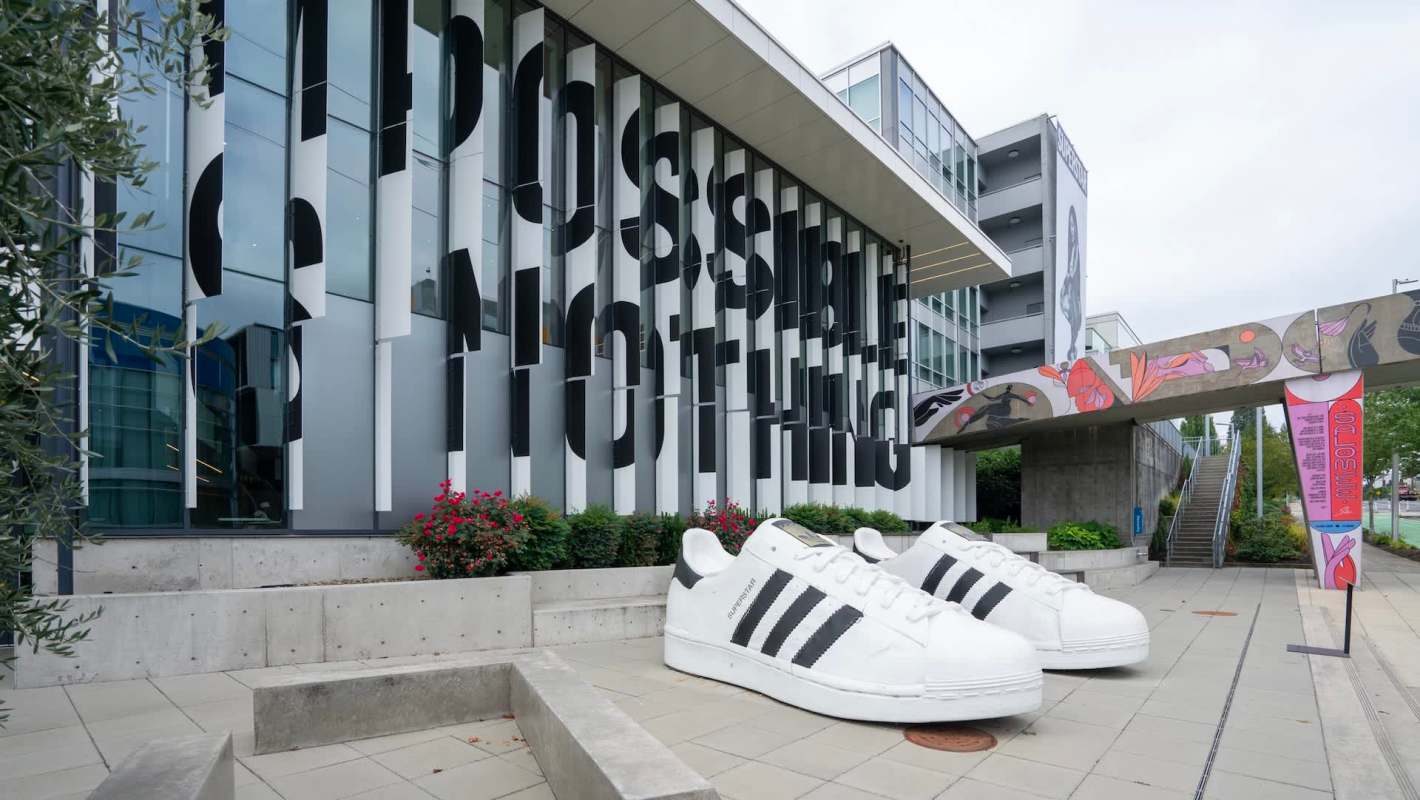 Oversized white Adidas sneakers with black stripes sit outside a modern building featuring "Impossible Nothing" signage.