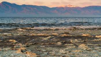 A rocky shoreline with salt formations and distant snow-capped mountains under a colorful sky.
