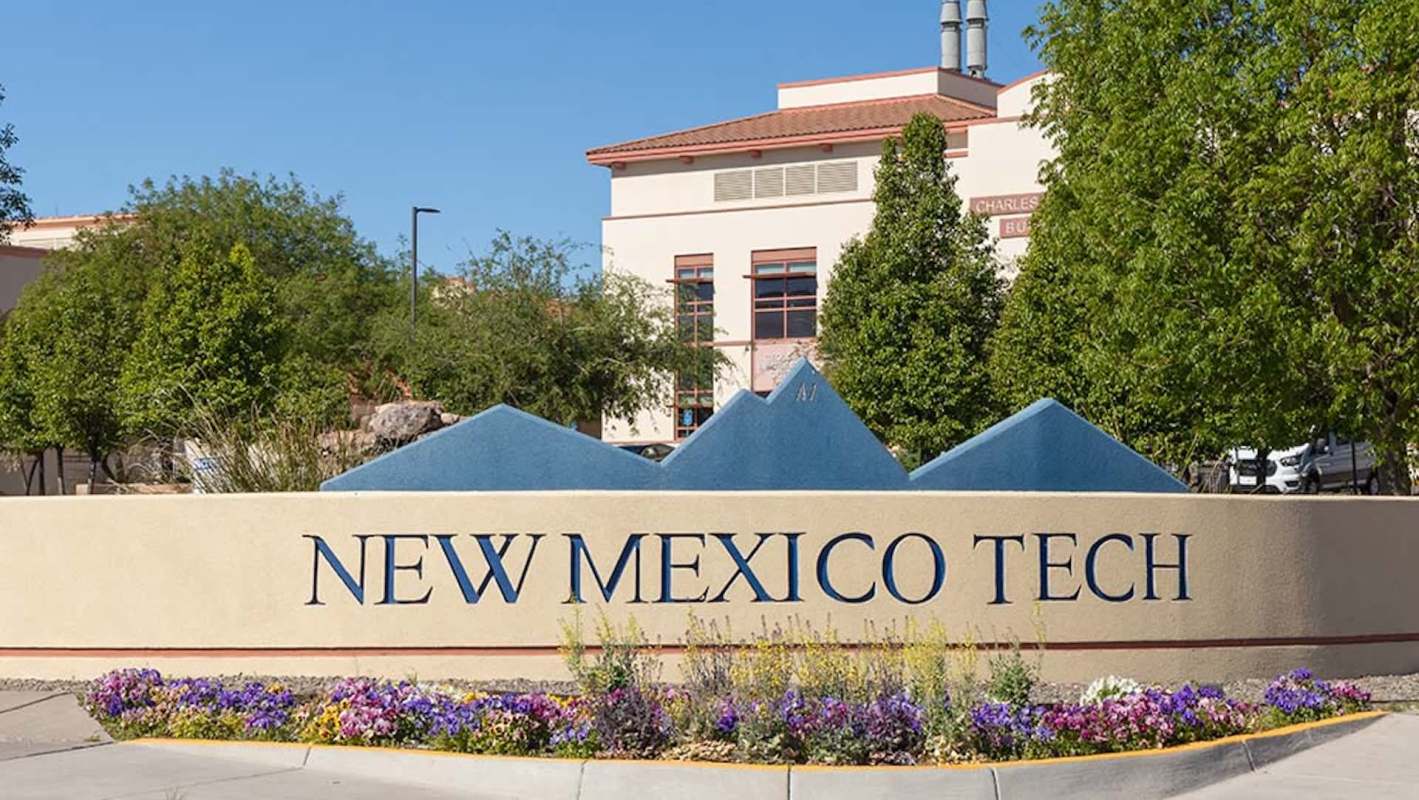 A sign for New Mexico Tech surrounded by colorful flowers and greenery under a clear blue sky.