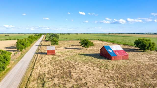 Aerial view of a Texas-themed barn surrounded by fields and a dirt road under a bright blue sky.