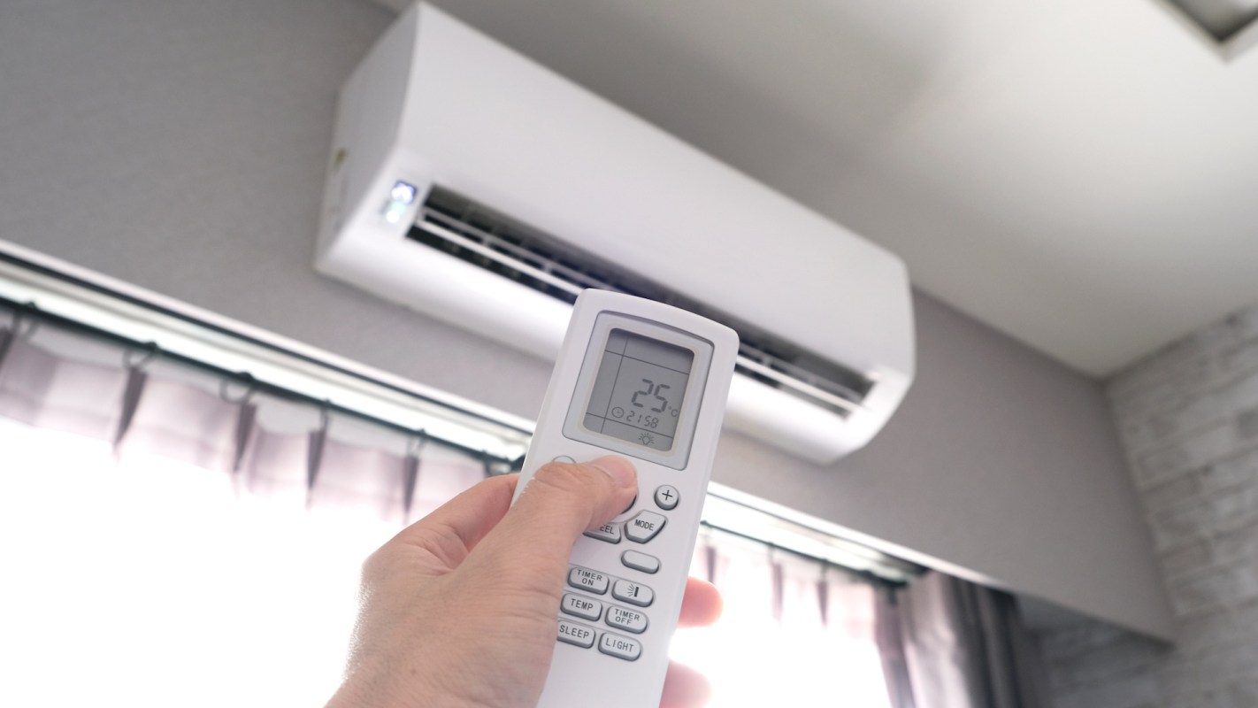 A person holds a remote control displaying the temperature setting for an air conditioning unit on the wall.