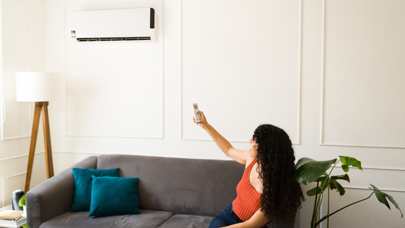 A woman sitting on a couch adjusts an HVAC with a remote.